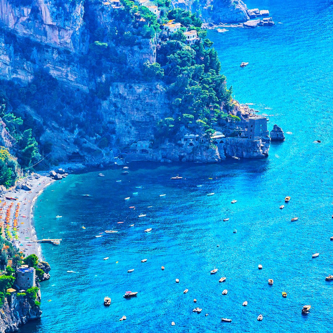 Veduta aerea di una cala riparata sulla Costiera Amalfitana con piccole barche da pesca all'ancora nell'acqua blu profondo e una spiaggia di ciottoli sotto le falesie calcaree