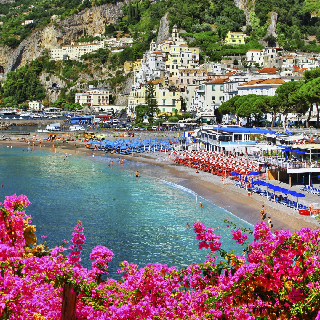 La spiaggia principale di Amalfi d'estate con bouganville in primo piano, ombrelloni a righe rosse e blu e la città medievale che sale sulla collina