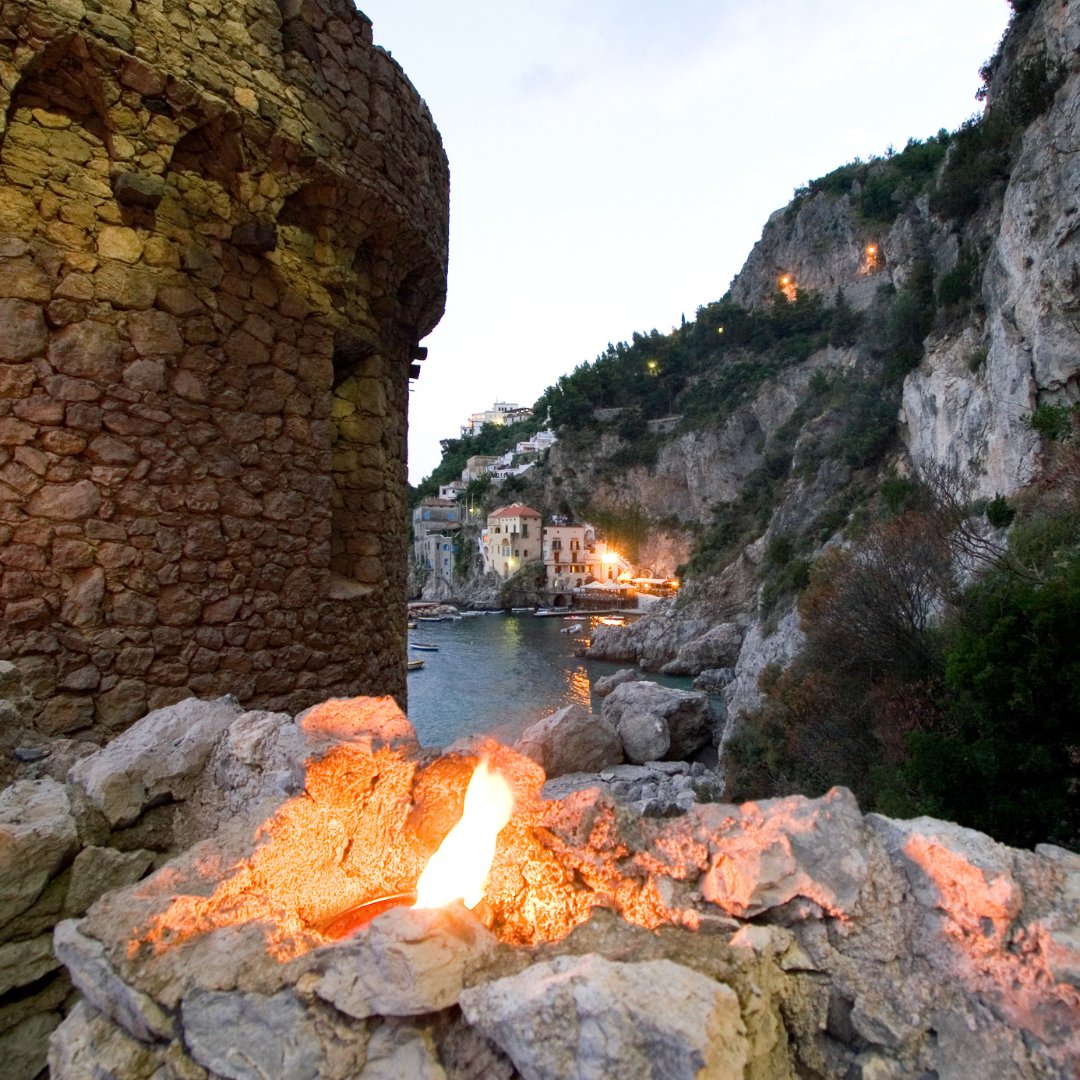 Una torre di avvistamento saracena medievale al tramonto sopra una cala riparata della Costiera Amalfitana, con un fuoco acceso in primo piano