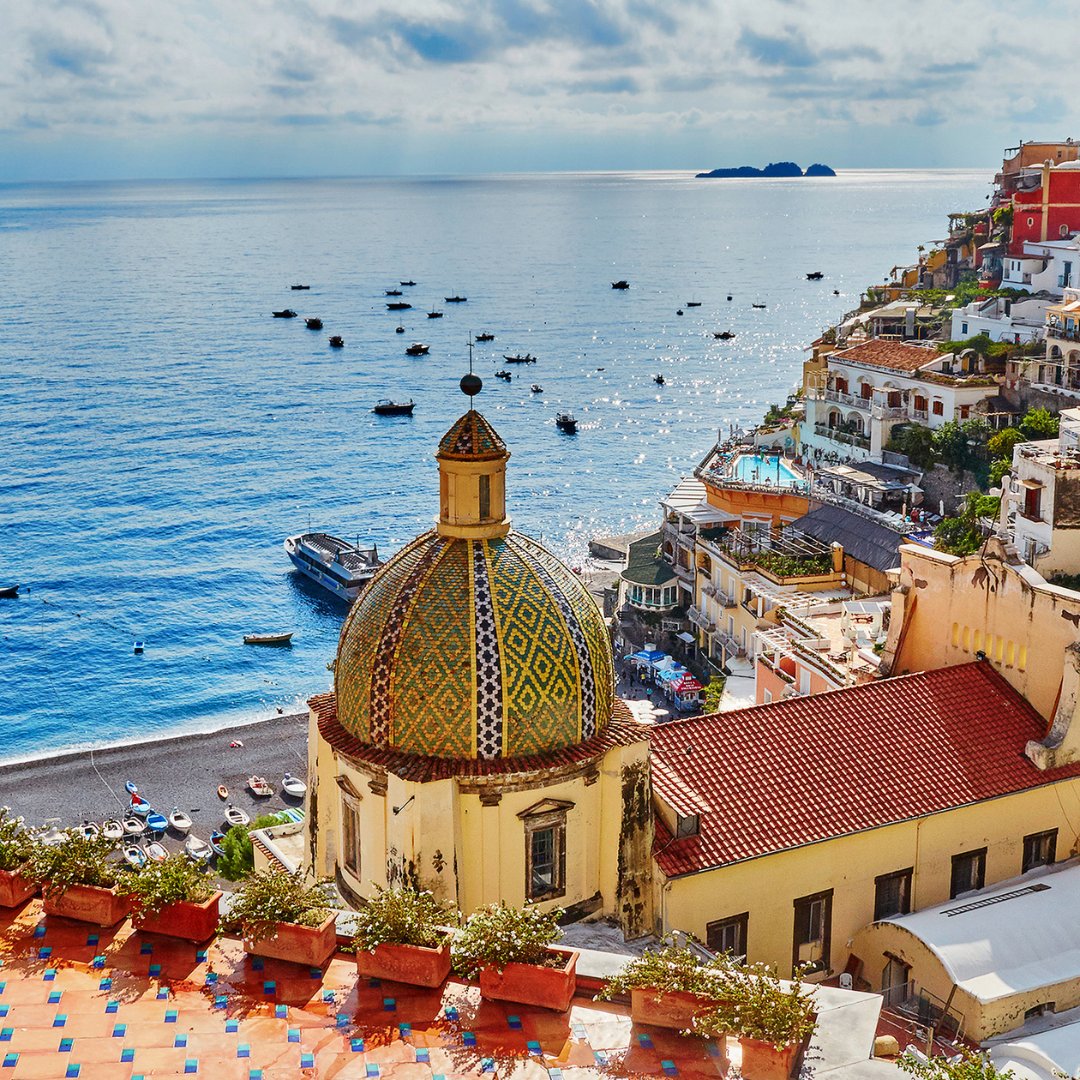 La cupola in maiolica di Santa Maria Assunta a Positano con i tetti in terracotta che scendono verso la spiaggia e le barche da pesca sul mar Tirreno