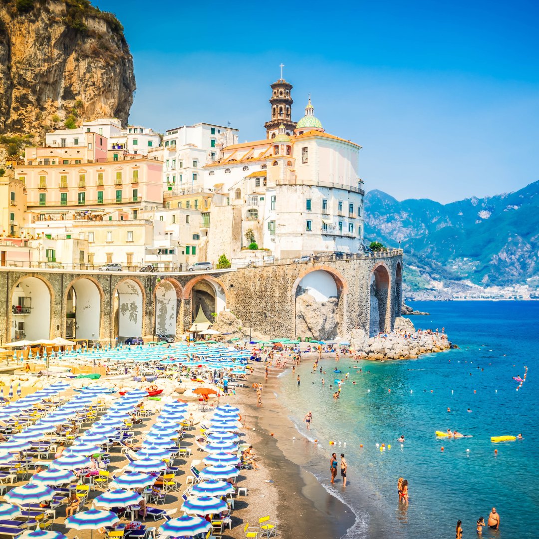 La spiaggia di Atrani d'estate con file di ombrelloni, bagnanti nell'acqua bassa e il campanile della Collegiata di Santa Maria Maddalena in alto