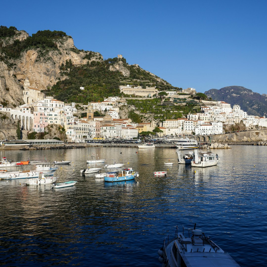 Il porto di Amalfi alla luce del mattino con le barche da pesca ormeggiate lungo il molo e la città medievale che si arrampica sulla collina, Campania, Italia meridionale